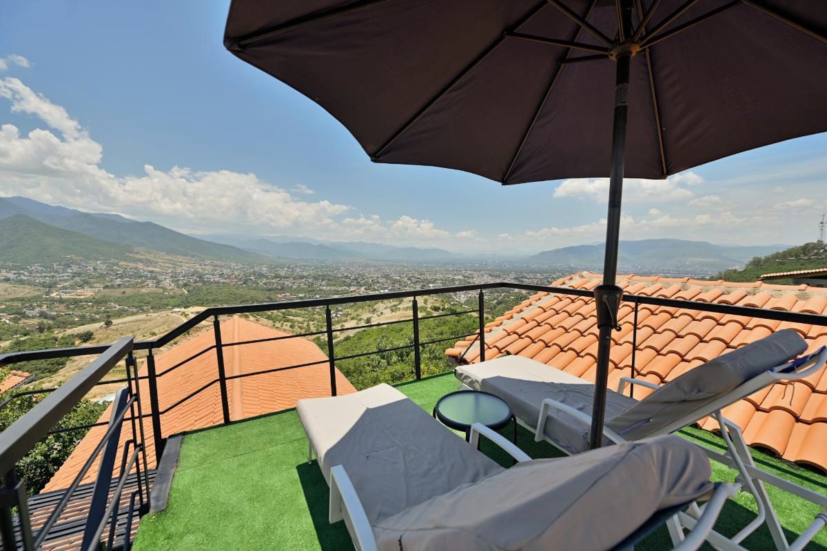 A balcony with a view of a cityscape and mountains, equipped with lounge chairs and a table.