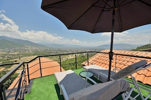A balcony with a view of a cityscape and mountains, equipped with lounge chairs and a table.
