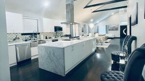 A modern kitchen with a central island, dark flooring, and a dining area with a black upholstered chair. - Elegant NW Atlanta Oasis, 2 Kitchens, Sauna, Billiards, Patio, Sundeck & Yard! (Atlanta)
