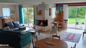 A cozy living room with a fireplace, a sofa, a dining table, and a view of a garden through large sliding doors.
