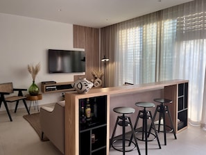 A modern living room with a wooden bar, a flat-screen TV, and a large window with sheer curtains.