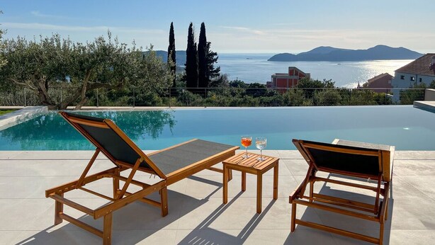 A poolside area with two wooden lounge chairs and a small table, overlooking a clear blue pool and a scenic view of the sea and mountains.