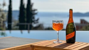A bottle of Monetto Roséco on a wooden table with a glass of rosé wine, overlooking a body of water and distant trees.