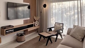 A modern living room with a flat-screen TV mounted on the wall, a wooden console table below, a black chair with a woven seat, a beige sofa, and a large window with sheer curtains.