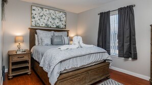 Ein Schlafzimmer mit einem hölzernen Bett, einem Nachttisch, einer Lampe, einem Fenster mit Vorhängen und einem floralen Bild an der Wand.