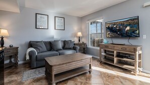 Ein Wohnzimmer mit grauem Sofa, Holztisch und Fernseher auf einem Holzständer. An den Wänden hängen zwei gerahmte Sprüche und ein Fenster mit Blick auf Häuser.