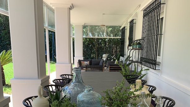 Una mesa de comedor dispuesta con diversos platos, vasos y adornos, rodeada de sillas y plantas en macetas, con vistas a un jardín y una zona de estar al aire libre.