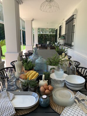 Ein Esstisch ist gedeckt mit verschiedenen Geschirr, Gläsern und Dekorationsartikeln, umgeben von Stühlen und Blumentöpfen, mit Blick auf einen Garten und eine Außenbereich-Sitzgelegenheit.