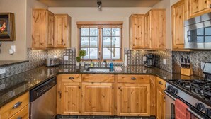 A kitchen with wooden cabinets, a stainless steel dishwasher, a stove, and a window with a view of snow outside. - Spacious Ski-In/Ski-Out Retreat (Big Sky)