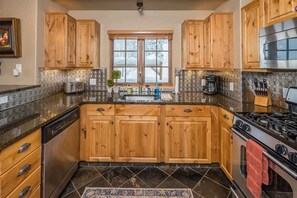 A kitchen with wooden cabinets, a stainless steel dishwasher, a stove, and a window with a view of snow outside. - Spacious Ski-In/Ski-Out Retreat (Big Sky)
