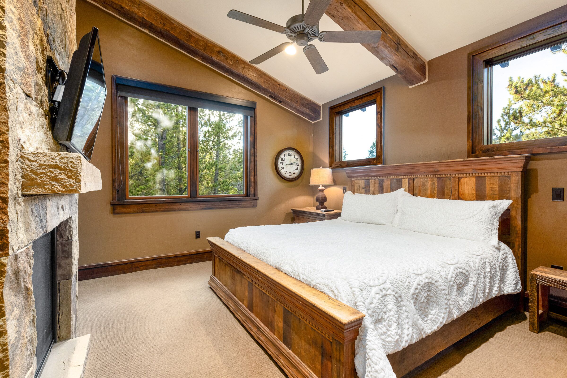 A bedroom with a wooden bed, stone fireplace, large windows, and a ceiling fan.