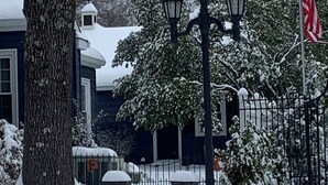 Un barrio residencial cubierto de nieve, con una casa, un árbol, una valla y un farol.