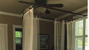 Una cama con dosel y cortinas traslúcidas, un ventilador de techo y un cuadro en la pared.