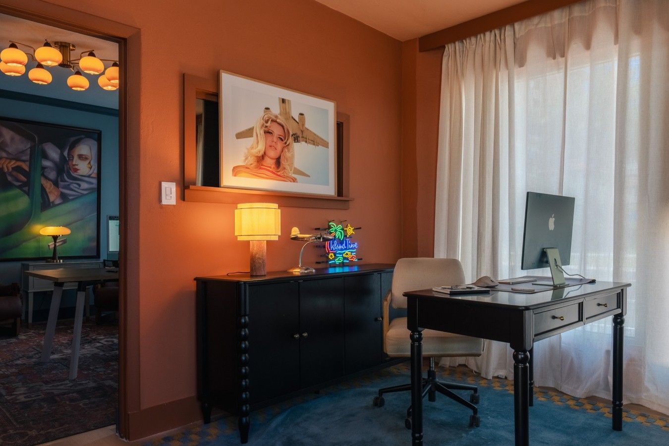 A hotel room with a desk, chair, and a large window with curtains. There is a painting on the wall and a lamp on the desk.