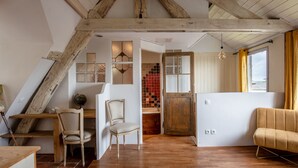 Une pièce avec un plafond en pente, des poutres en bois, un bureau en bois, une chaise, un canapé et une vue sur un espace carrelé, visible à travers une porte.