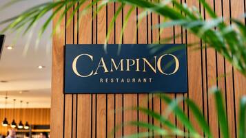 A sign for 'Campino Restaurant' with a wooden backdrop and green foliage in the foreground.