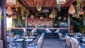 An outdoor dining area with wicker chairs and tables, a bar with bottles, and hanging plants. - Longue Vie Hôtels (Marrakech)
