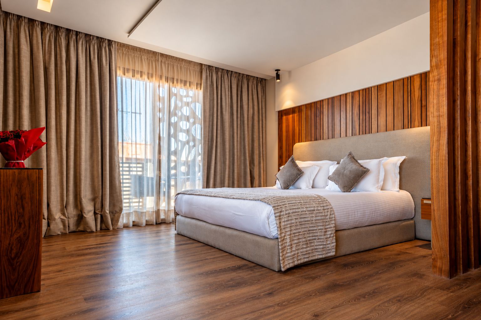 Un dormitorio moderno con una cama grande, paneles de madera en las paredes y vistas al exterior a través de cortinas traslúcidas.