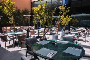 An outdoor seating area with round tables and wicker chairs, surrounded by potted plants and a modern building facade.