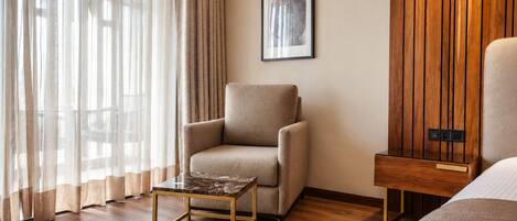 A hotel room with a wooden floor, a beige armchair, a small table, a bed, and a wooden panel wall.