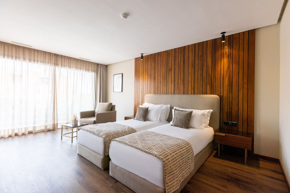 Habitación de hotel con dos camas, una pared decorativa de madera, un ventanal con cortinas translúcidas, una mesita y una silla.