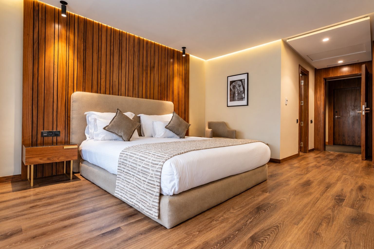A modern bedroom with a large bed, wooden paneling on the wall, a bedside table, and a door.