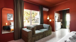A room with a green sofa, a bed, a window with curtains, a mirror, and a kitchenette.