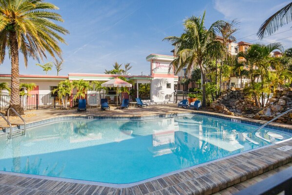 A swimming pool with lounge chairs and umbrellas, surrounded by palm trees and a building with a sign.