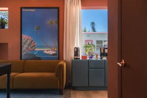 Une pièce comprenant un canapé, un tableau, un bureau avec un ordinateur et une vue sur un immeuble par la fenêtre.