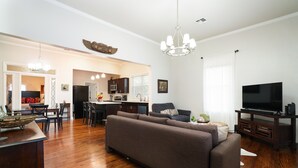 Uma sala de estar moderna com um sofá seccional, uma área de jantar com cadeiras, uma cozinha ao fundo e uma TV de tela plana sobre um suporte.