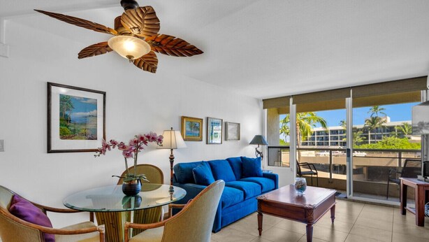 Ein Wohnzimmer mit blauem Sofa, einem Glastisch, Korbstühlen, einem Deckenventilator und einem Balkon mit Blick auf Palmen.