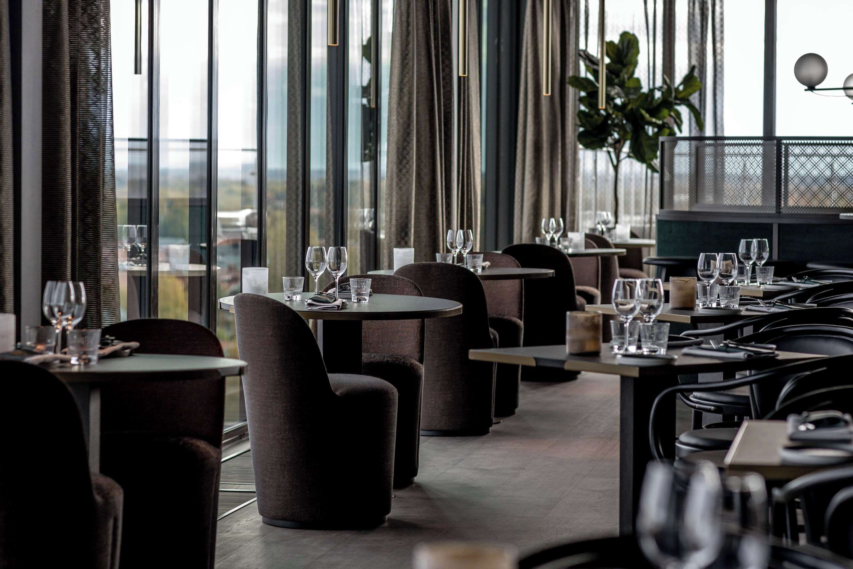 Un restaurante con mesas redondas y sillas oscuras, ventanas amplias con cortinas y vistas al exterior.