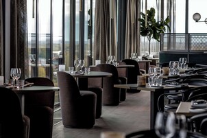 Un restaurante con mesas redondas y sillas oscuras, ventanas amplias con cortinas y vistas al exterior.