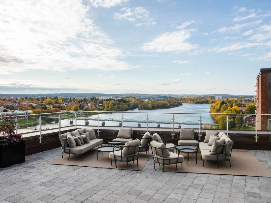 A balcony with outdoor seating, a view of a river, and a cityscape in the background.