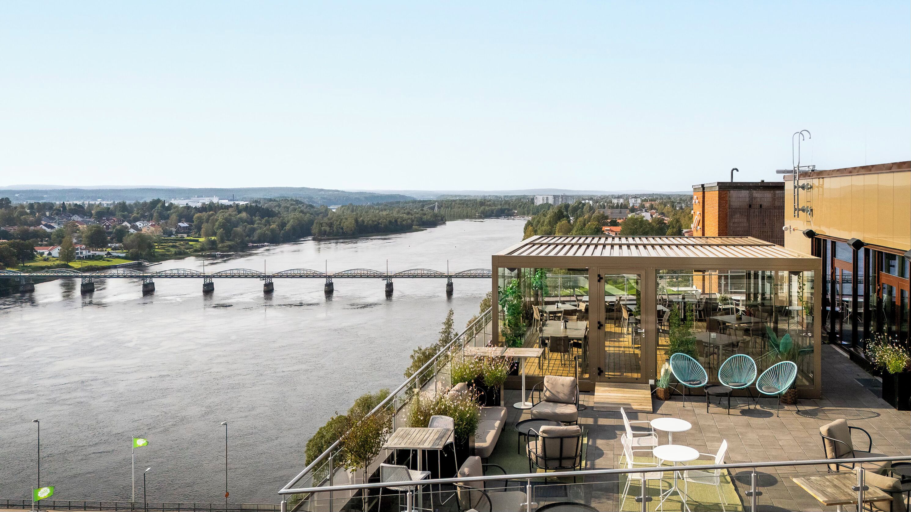 A riverside terrace with outdoor seating, a bridge in the background, and a view of a residential area.