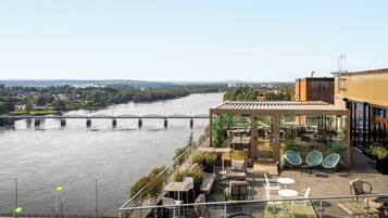 A riverside terrace with outdoor seating, a bridge in the background, and a view of a residential area.
