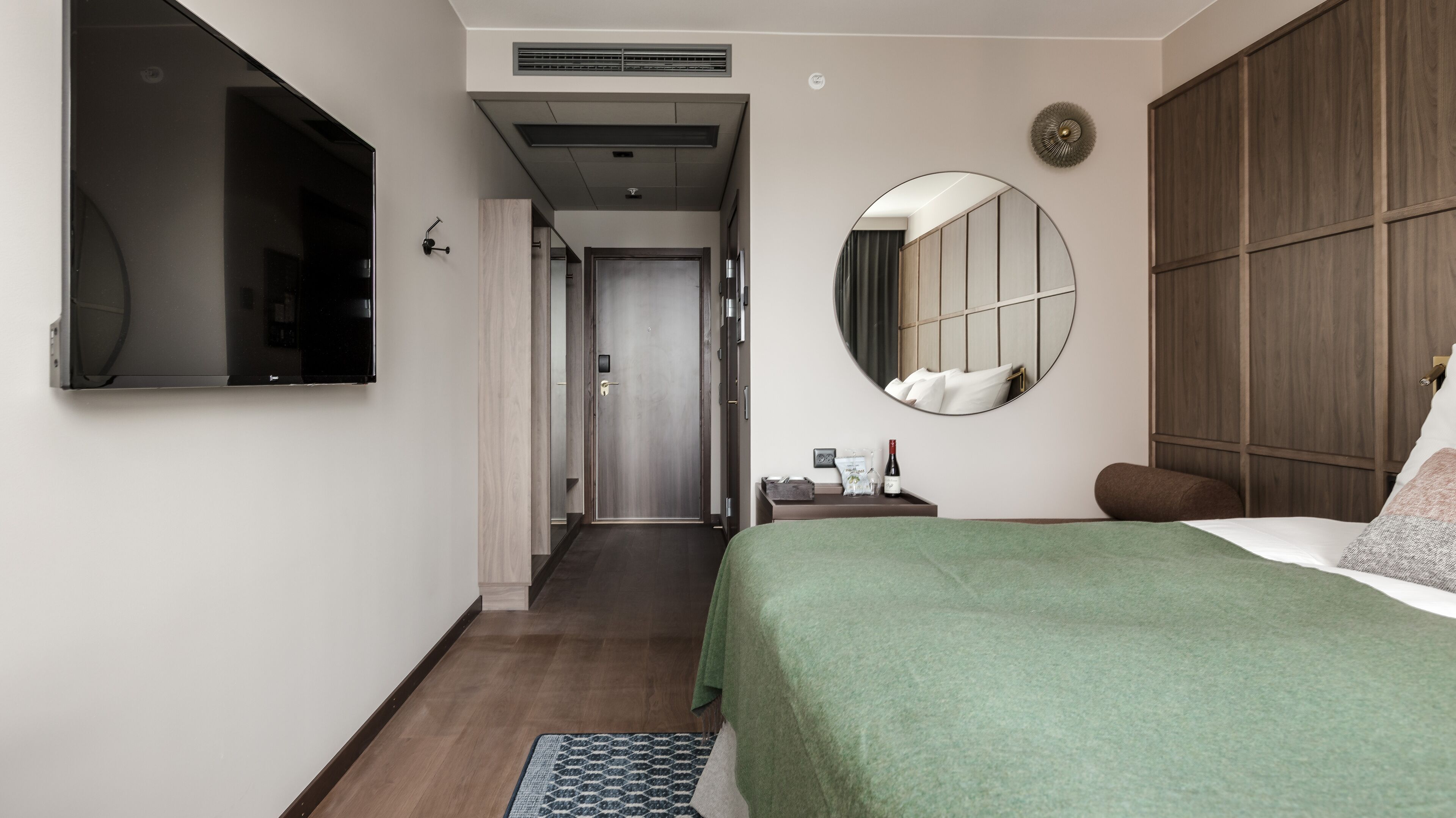 Quarto de hotel com cama, mesa de cabeceira, espelho e armário. Há um corredor que leva a outro quarto.