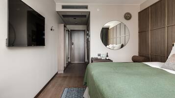 Quarto de hotel com cama, mesa de cabeceira, espelho e armário. Há um corredor que leva a outro quarto.