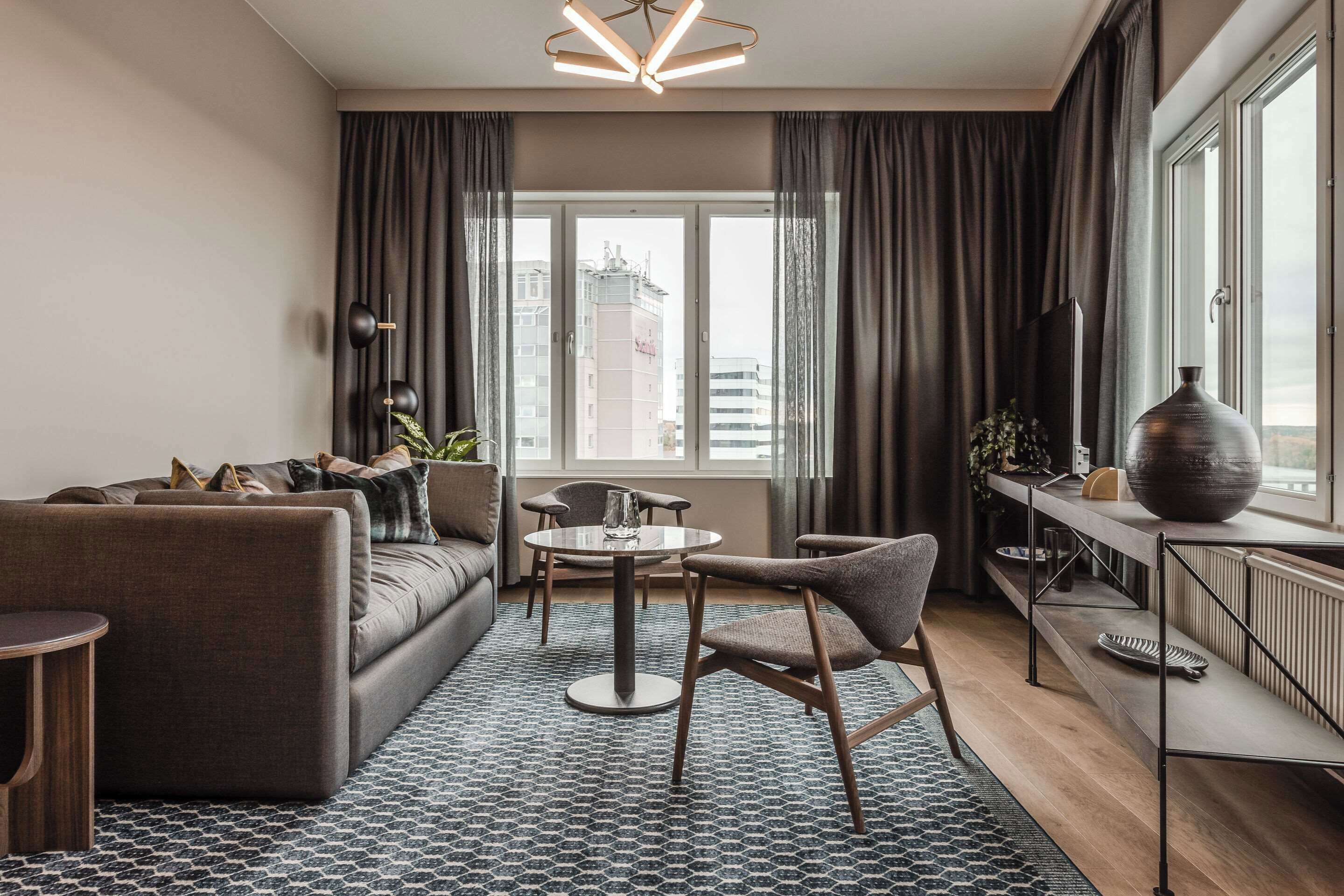 Un salon moderne avec un canapé, un fauteuil et une table ronde. On y trouve une grande fenêtre avec des rideaux, un radiateur et un vase posé sur une étagère.