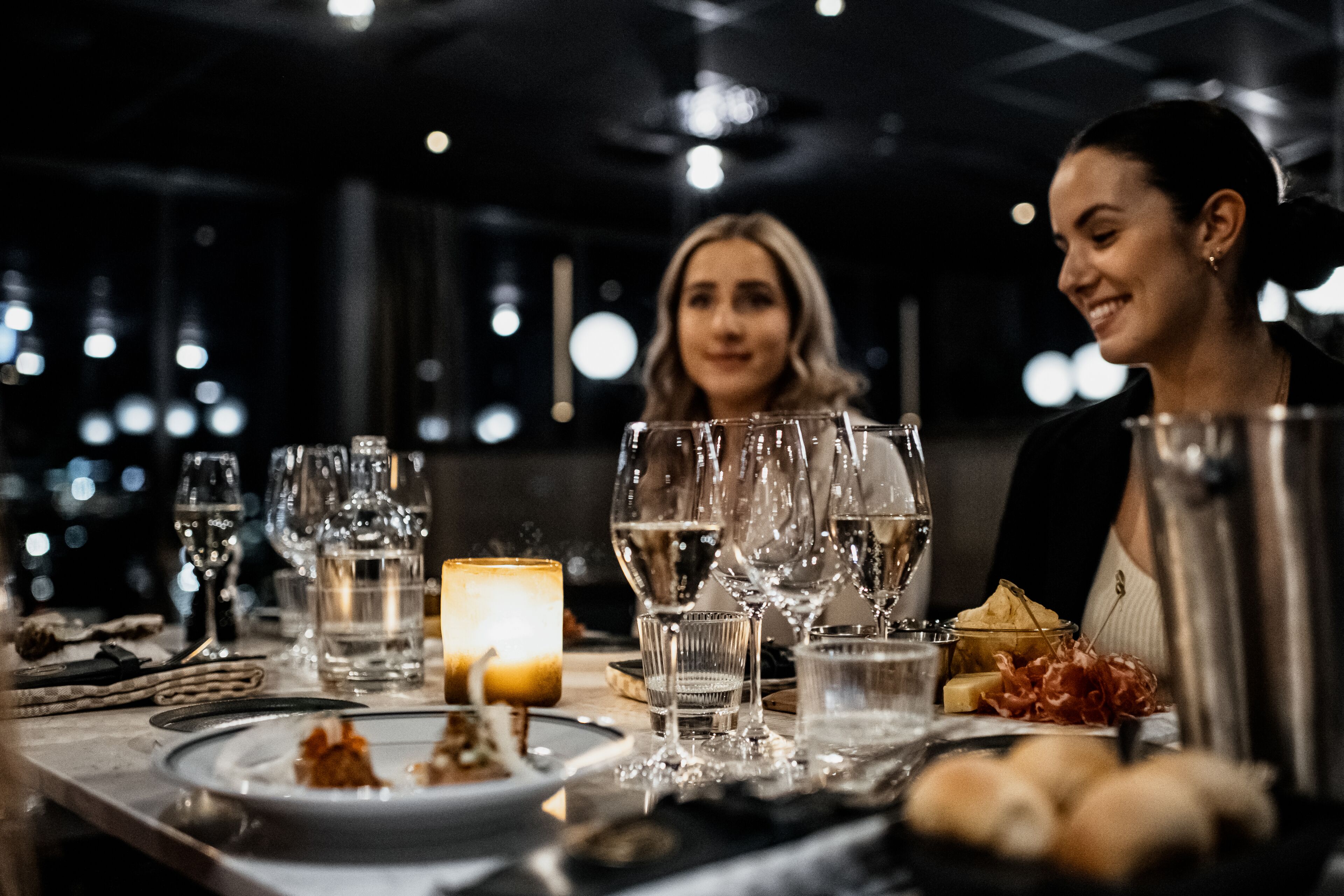 A dimly lit restaurant interior with a table set for two, featuring wine glasses, a candle, and appetizers.