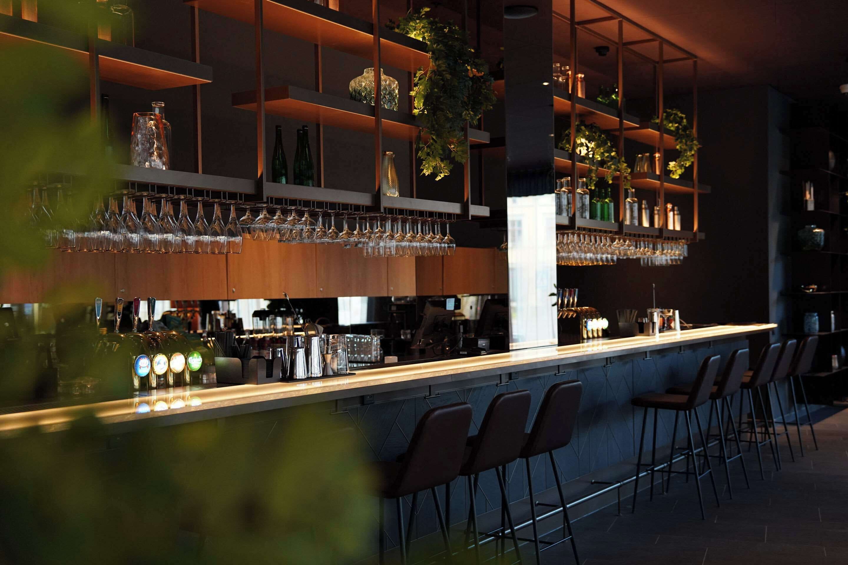 Una zona de bar con estantes llenos de botellas y vasos, una barra con utensilios y taburetes de bar.