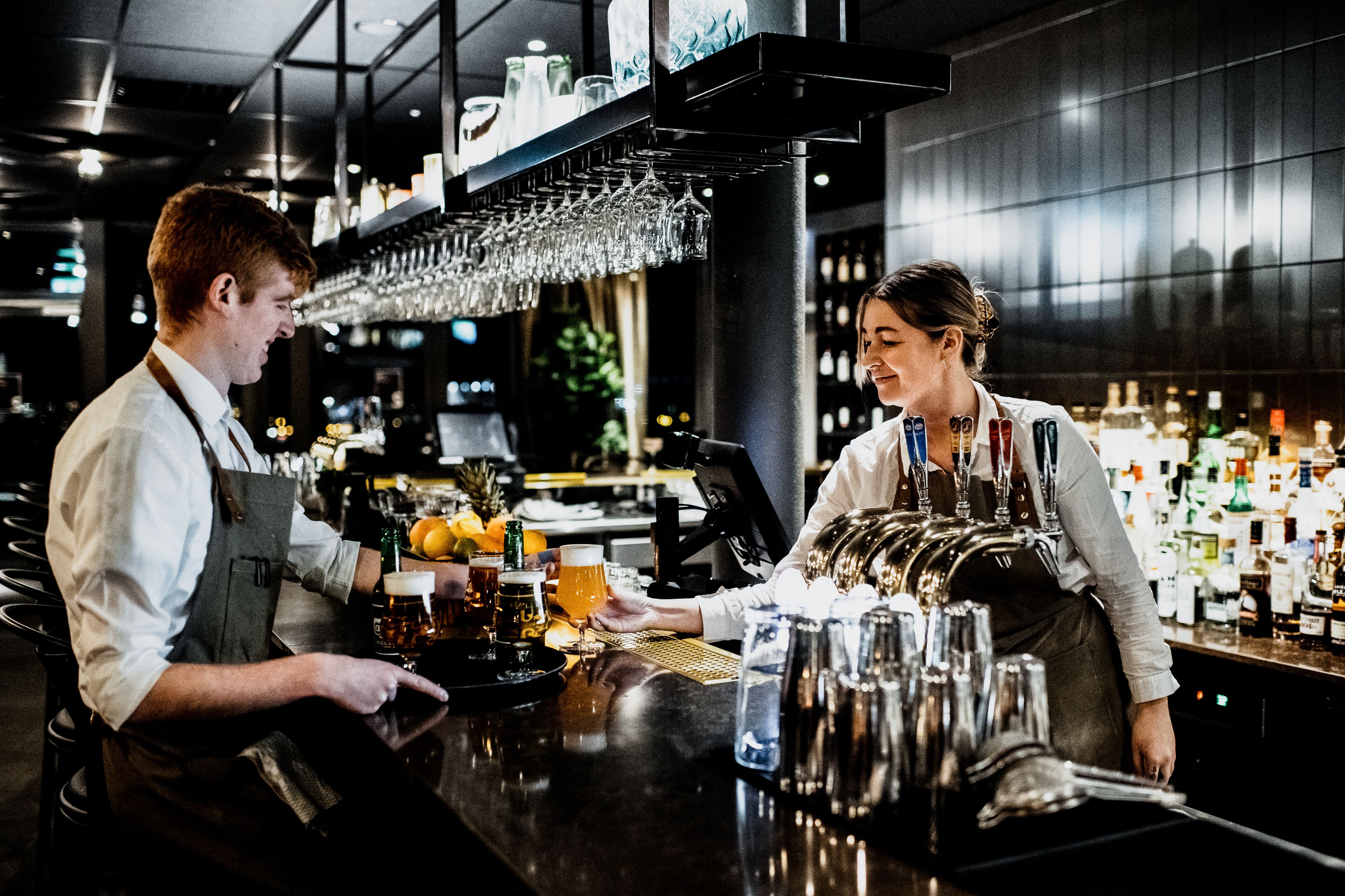 Un bar avec un barman et un client. Le bar présente un design moderne, avec un comptoir noir et une variété de bouteilles et de verres.