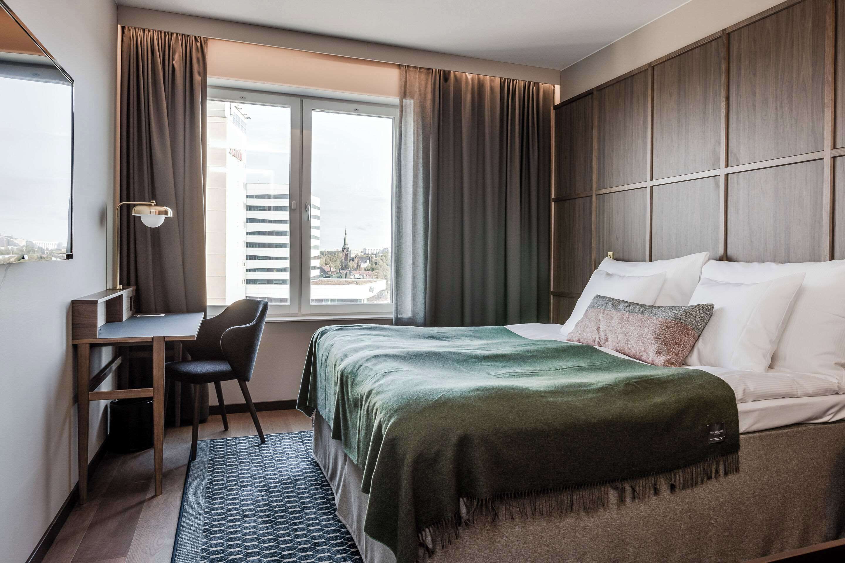 A hotel room with a bed, desk, chair, and a view of the city through a window.