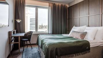 A hotel room with a bed, desk, chair, and a view of the city through a window.