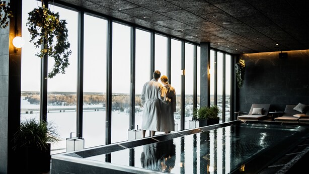 A couple standing by a large indoor pool with a view of a snowy landscape through large windows. - Clarion Hotel Umea (Umea)