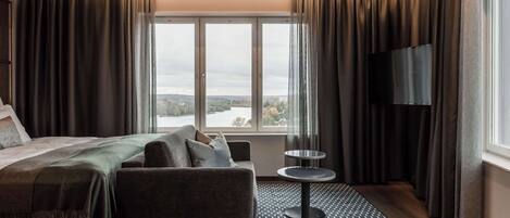 Habitación de hotel con cama, sofá, una mesita, televisión y ventana con cortinas.