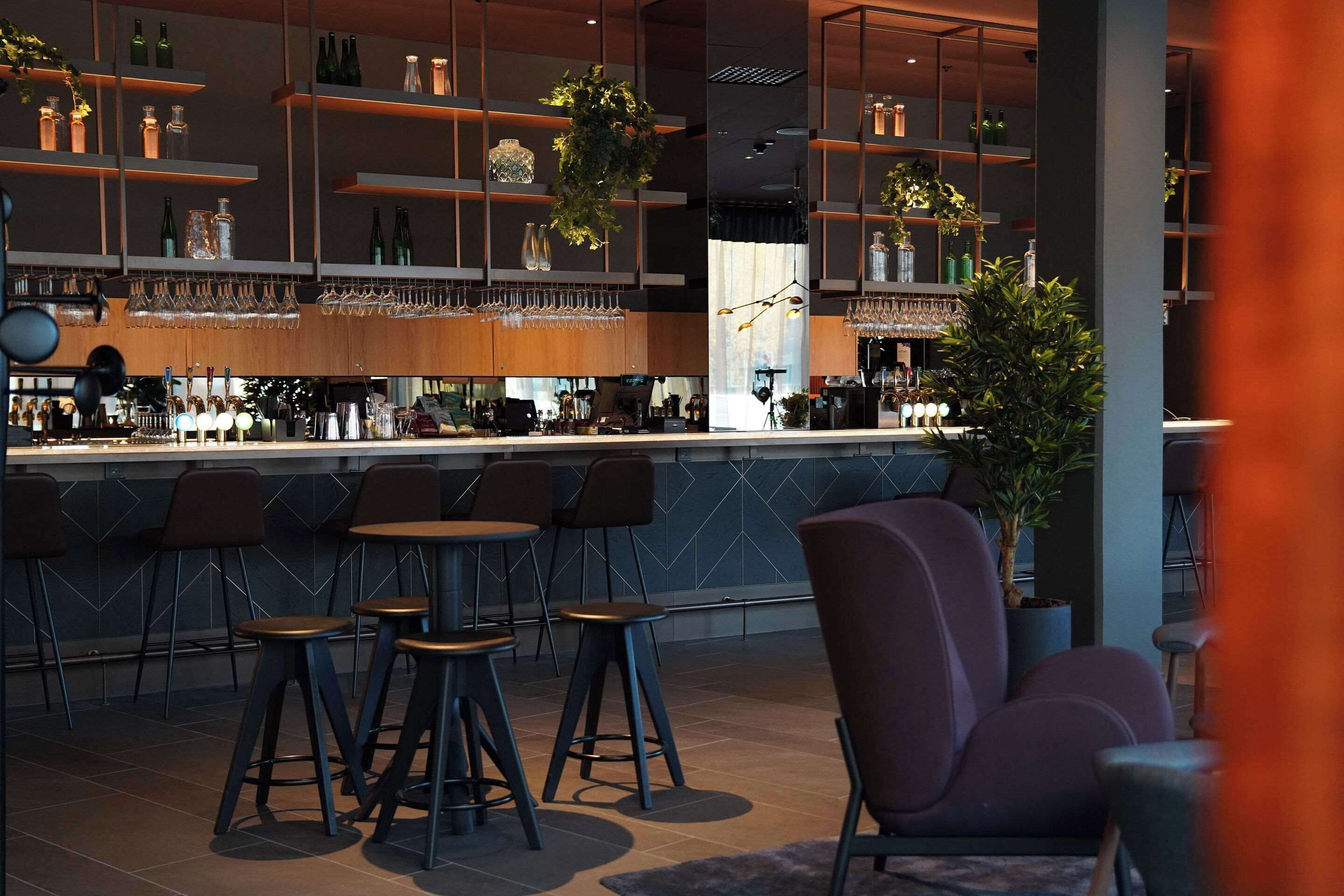 A modern bar area with multiple stools, shelves with bottles and glasses, and a potted plant.