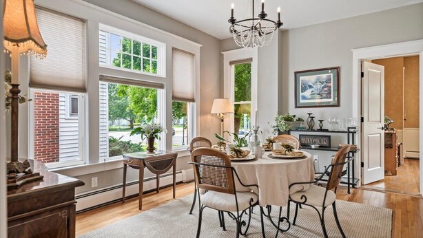 A dining area with a round table, chairs, and a chandelier. There are windows with blinds, a brick wall, and a door leading to another room.