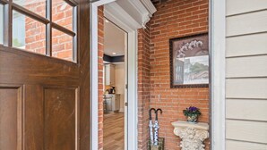 A wooden door leading to a room with a brick wall, a framed picture, and a small table with a potted plant.