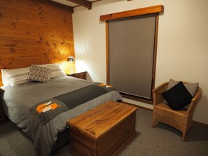 Een slaapkamer met een houten hoofdbord, een bed met grijze beddengoed, een nachtkastje van hout met een lamp, een rieten stoel en een houten dressoir.
